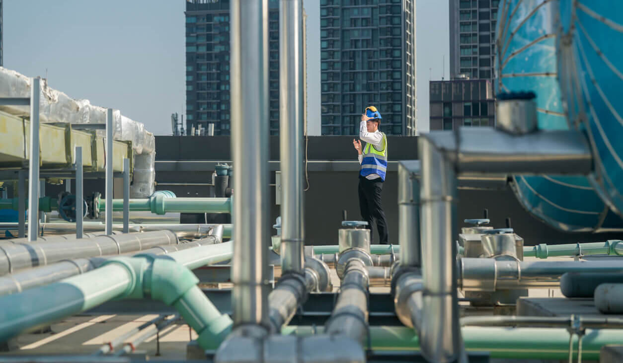 A professional engineer communicates via walkie-talkie on a building's rooftop, surrounded by piping and ductwork.